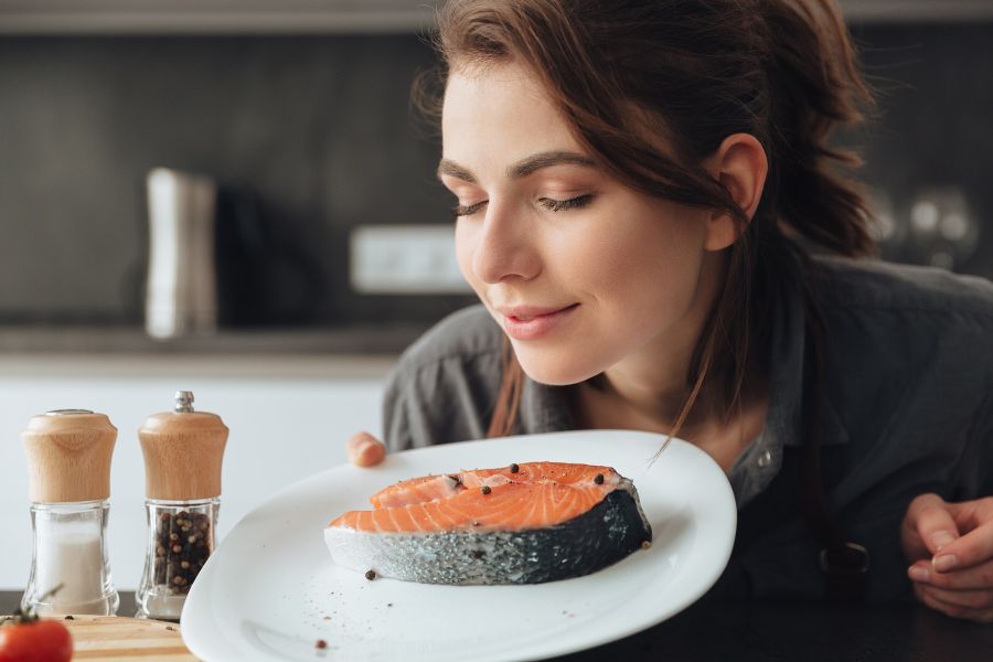 Photo of young pretty woman standing in kitchen while cooking and smells fish. Eyes closed.
